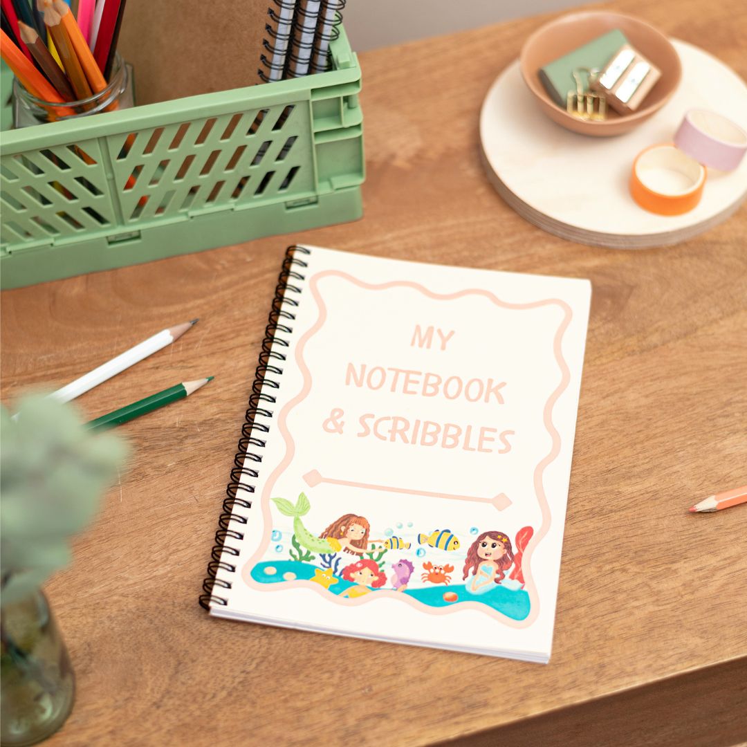 A5 Kids Mermaid Notes and Scribbles Notebook. A Handmade A5 Notebook with a mermaid theme placed on a wooden desk alongside stationery items.