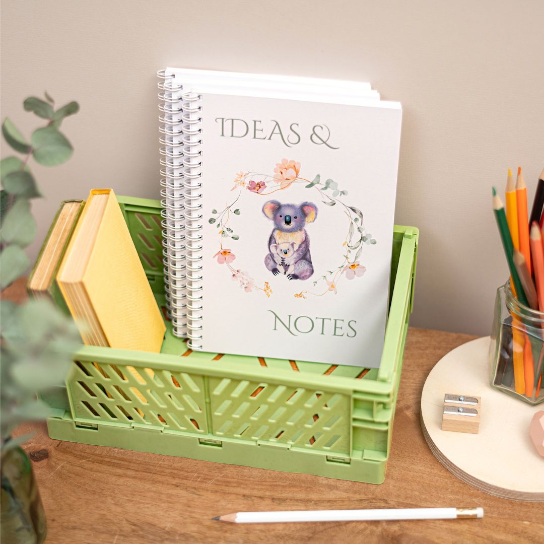 A5 Koala Wirebound Notebook. A handmade A5 notebook with a koala and baby design on the cover, titled 'Ideas & Notes'. It is placed on a desk next to a pen, pencils, and a plant.