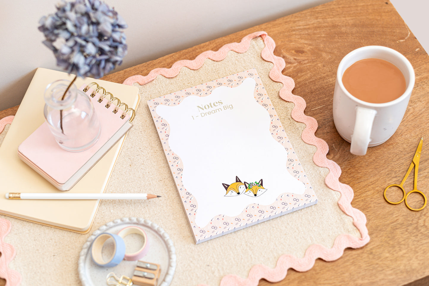 An A5 daily planner pad with a floral and fox design on the cover, placed on a wooden desk next to a cup of tea, a notebook, pens, and scissors.