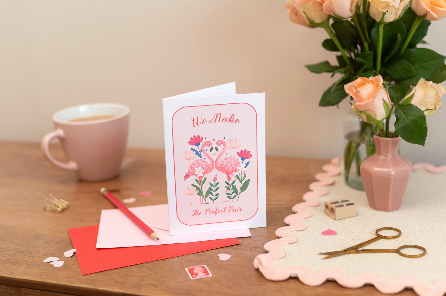 A Valentines Romance Friendshop greeting card with a flamingo design, along with an envelope and a pair of scissors, placed on a table with a vase of pink roses in the background. The text says "we make the perfect pair"