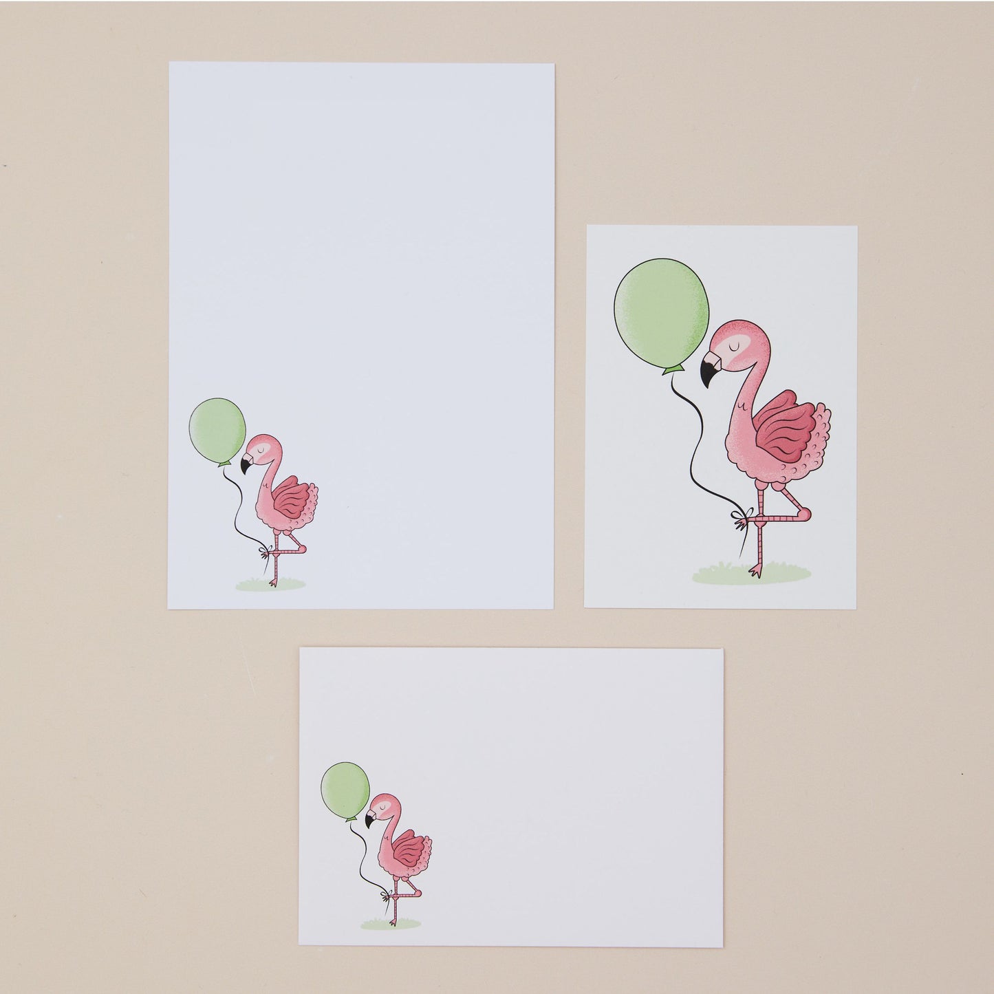 Pink Flamingos Writing Set Illustrations. Three illustrations of pink flamingos with green balloons on a beige background. One shows the writing paper, another shows the postcard, and another the front of the envelope all featuring the pink flamingos and green balloons