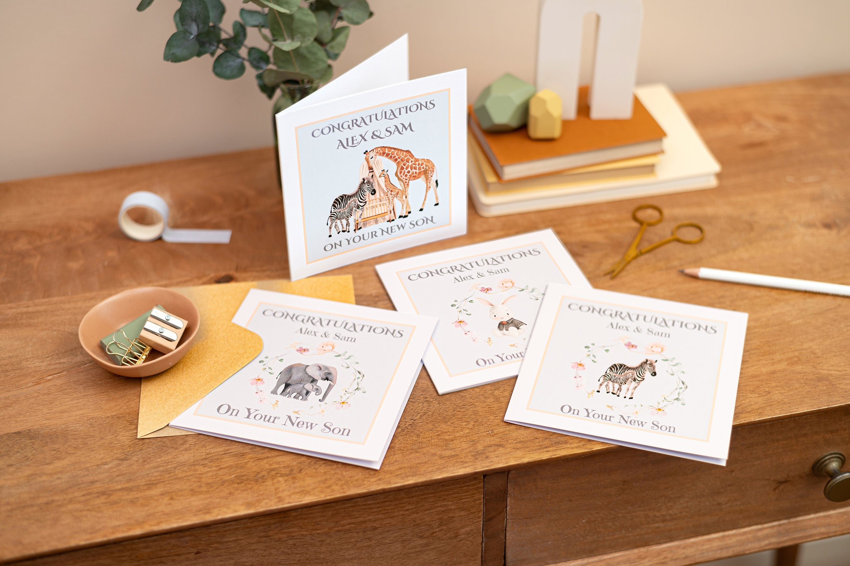 New Baby Cards. New Baby Boy, New Baby Girl, New Grandparents Congratulations Cards, Mummy to be  Cards. Illustrations of Elephants, Giraffes, Zebras, Rabbits with their young. The cards are in a group on a table with books, stationery and dried flowers