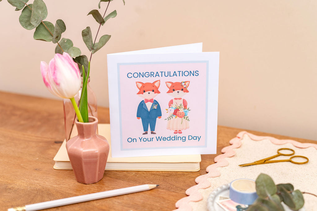 Anniversary Cards. A Group of Anniversary Congratulations cards featuring cartoon fox couples. Cards are on a wooden desk with books, candles, foliage and pink glitter ball