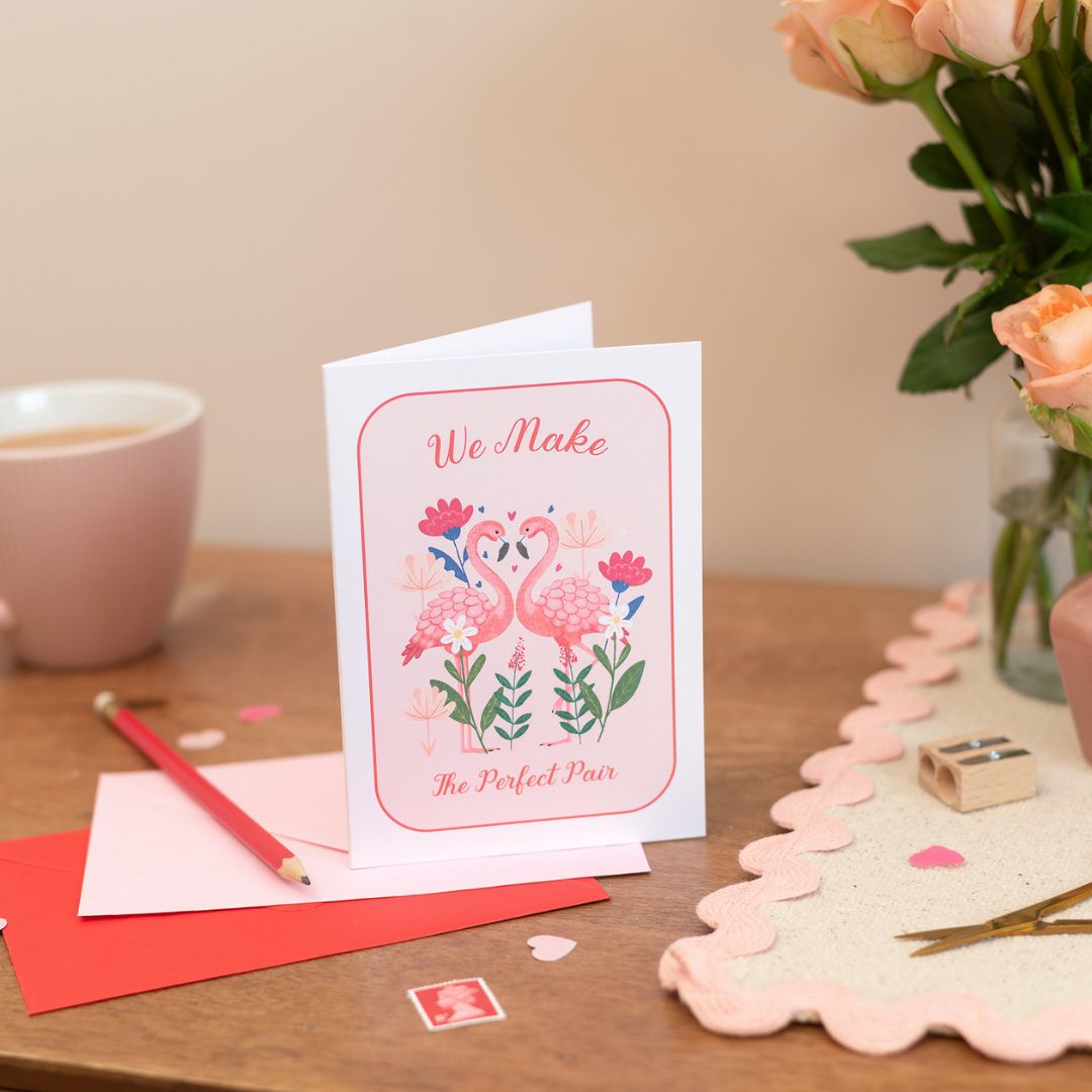 Romance and Friendship Card. A6 Greeting Card with the text "we make the perfect pair". The card features a pair of flamingos set in a romantic setitng.There are pink and red envelopes on the table along with Roses and stationery