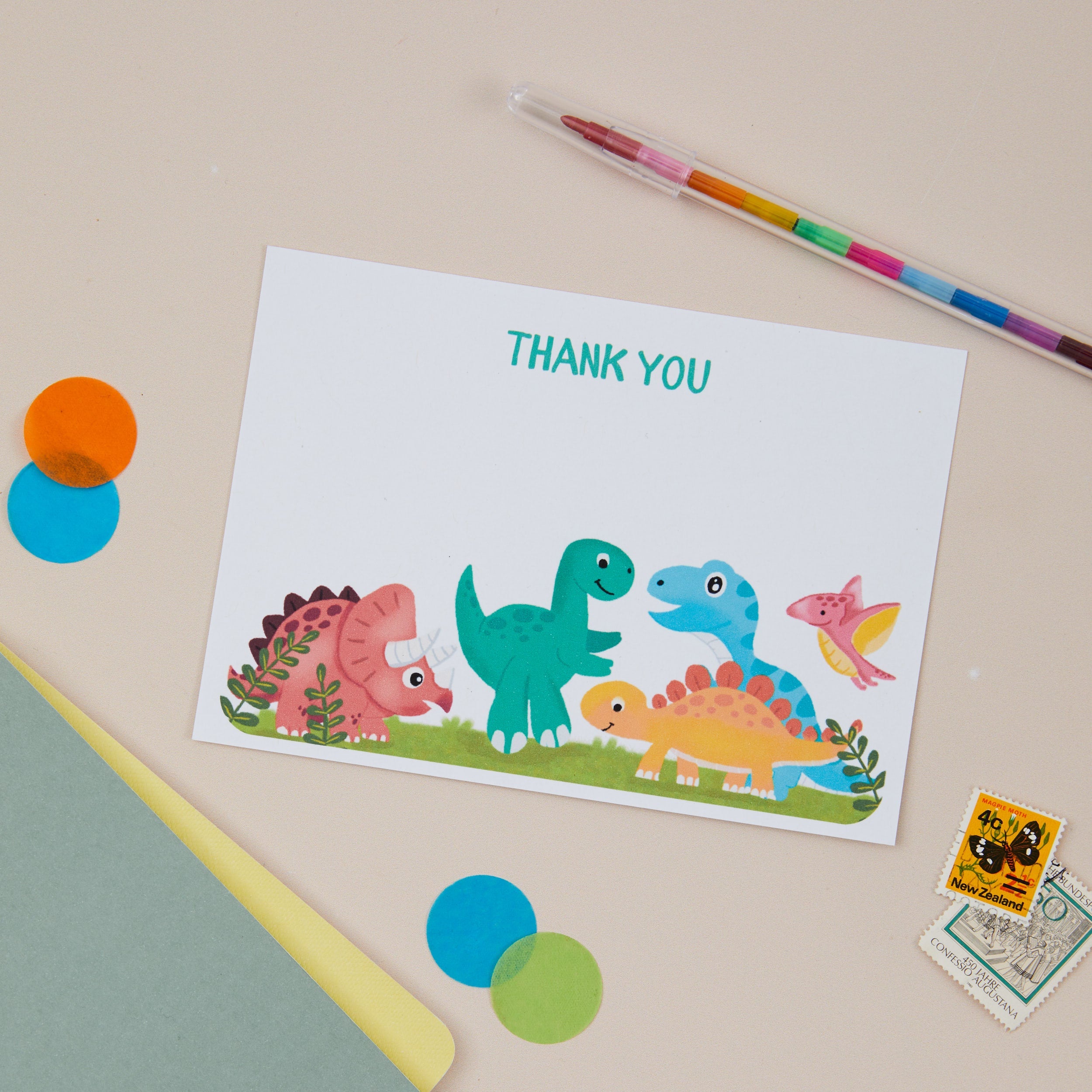 Children's Postcards. A6 Children's Postcard which has an illustration of a set of Baby Dinosaurs. The Dinosaurs are on grass. The Kids Postcard sits on a beige background and stationery surrounds it