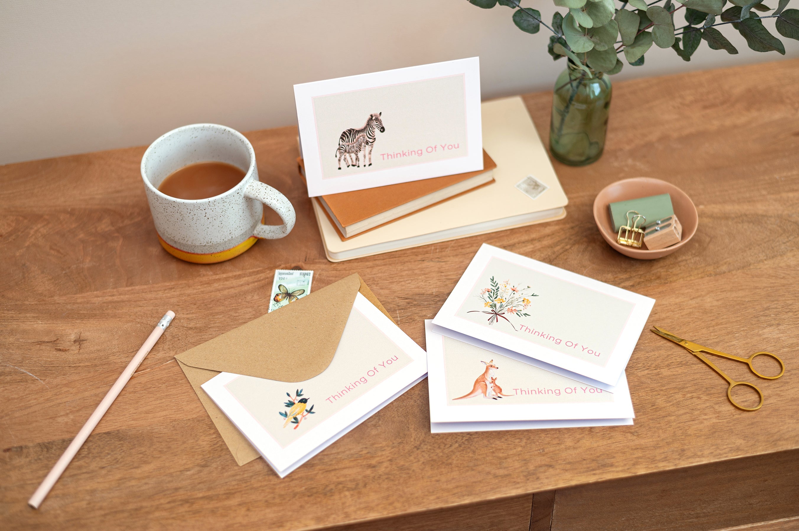 Thinking Of You Cards. Group photo of illustrations of Thinking of you cards. The cards feature a zebra, bird, kangaroo & floral bouquet. The cards rest on a wooden table with a cup of tea