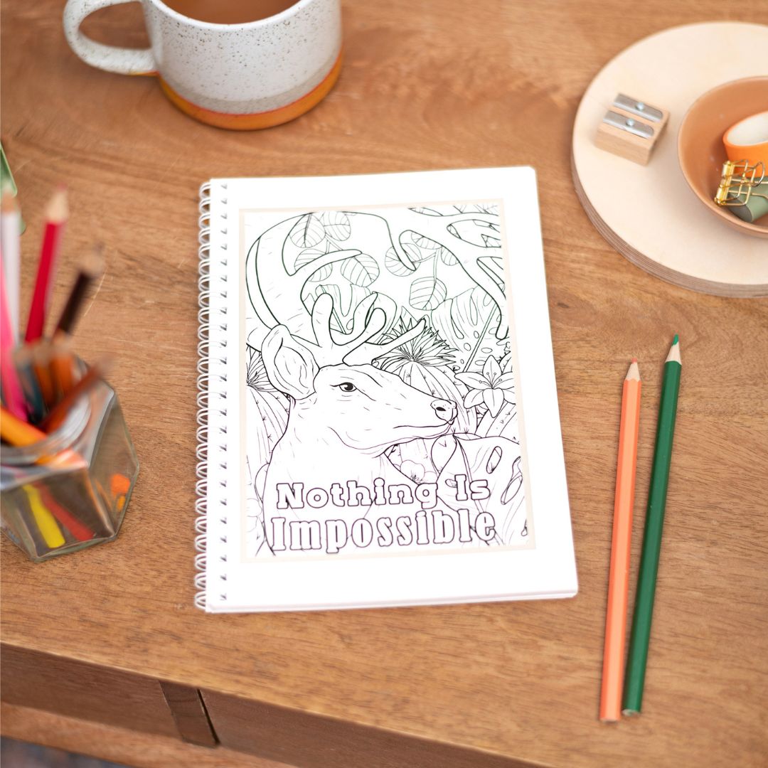A5 Colouring in Nothing is Impossible Notebook. A hand-drawn, motivational notebook with the phrase 'Nothing Is Impossible' on the cover, placed on a wooden desk alongside a cup of coffee, pencils, and a notebook.