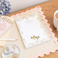 An A5 daily planner pad with a mosaic-inspired design and a 'Dream Big' reminder on the cover, displayed on a wooden desk alongside a cup of tea, a notepad, a pair of scissors, and a decorative element.