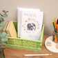 A handmade spiral-bound notebook with an elephant and floral design on the cover, titled 'Ideas & Notes'. It is placed inside a green plastic crate on a wooden desk alongside books, pencils, and a plant.
