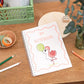 A5 Kids Flamingo Notebook. A handmade A5 notebook with a white cover featuring a pink flamingo and greenery design, placed on a wooden table alongside pencils and a plant.