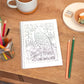An A5 motivational notebook with a bird illustration on the cover, titled 'Find Your Balance', is placed on a wooden table alongside a cup of coffee, a sharpener, and a set of colored pencils.