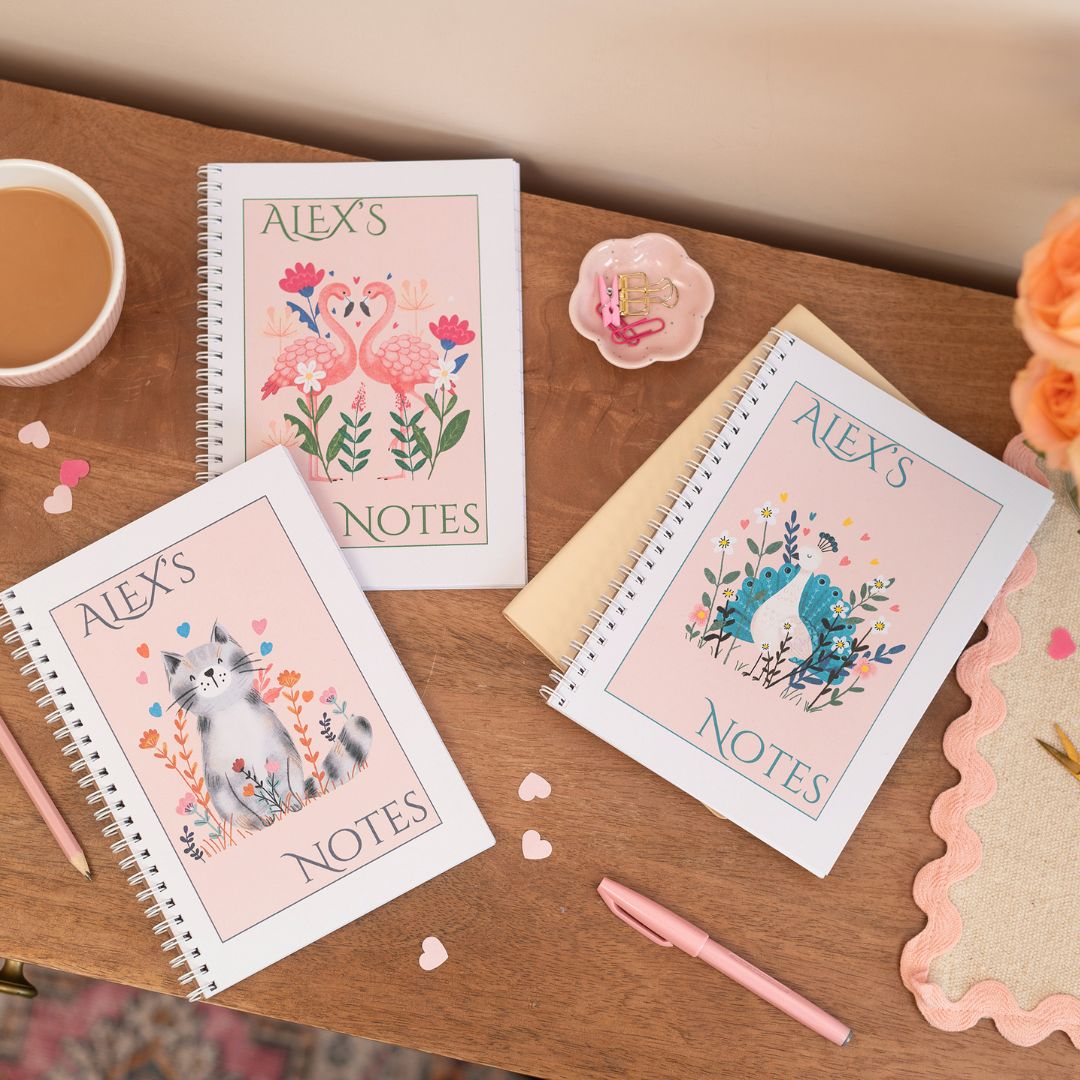 Group of 3 Personalised A5 Notebooks which are wire bound. The notebooks have animal characters on the front against a pink background. They are on a wooden table with a cup of coffee, flowers, and stationery items.