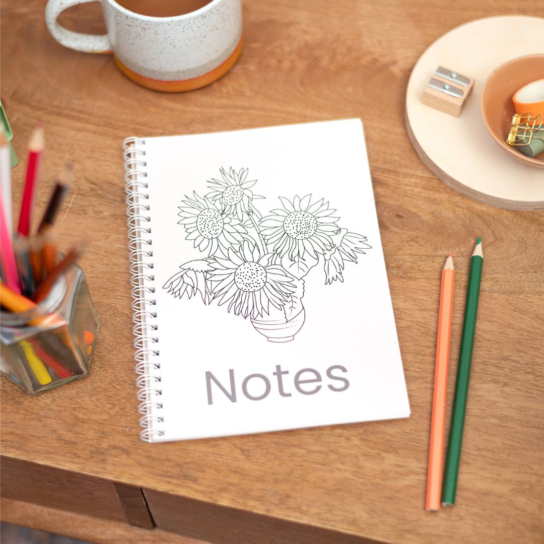 A5 Sunflowers Colouring In Notebook. An A5 notebook with a colouring cover of Sunflowers, titled 'Notes', placed on a wooden table alongside a cup of coffee, a sharpener, and coloured pencils.