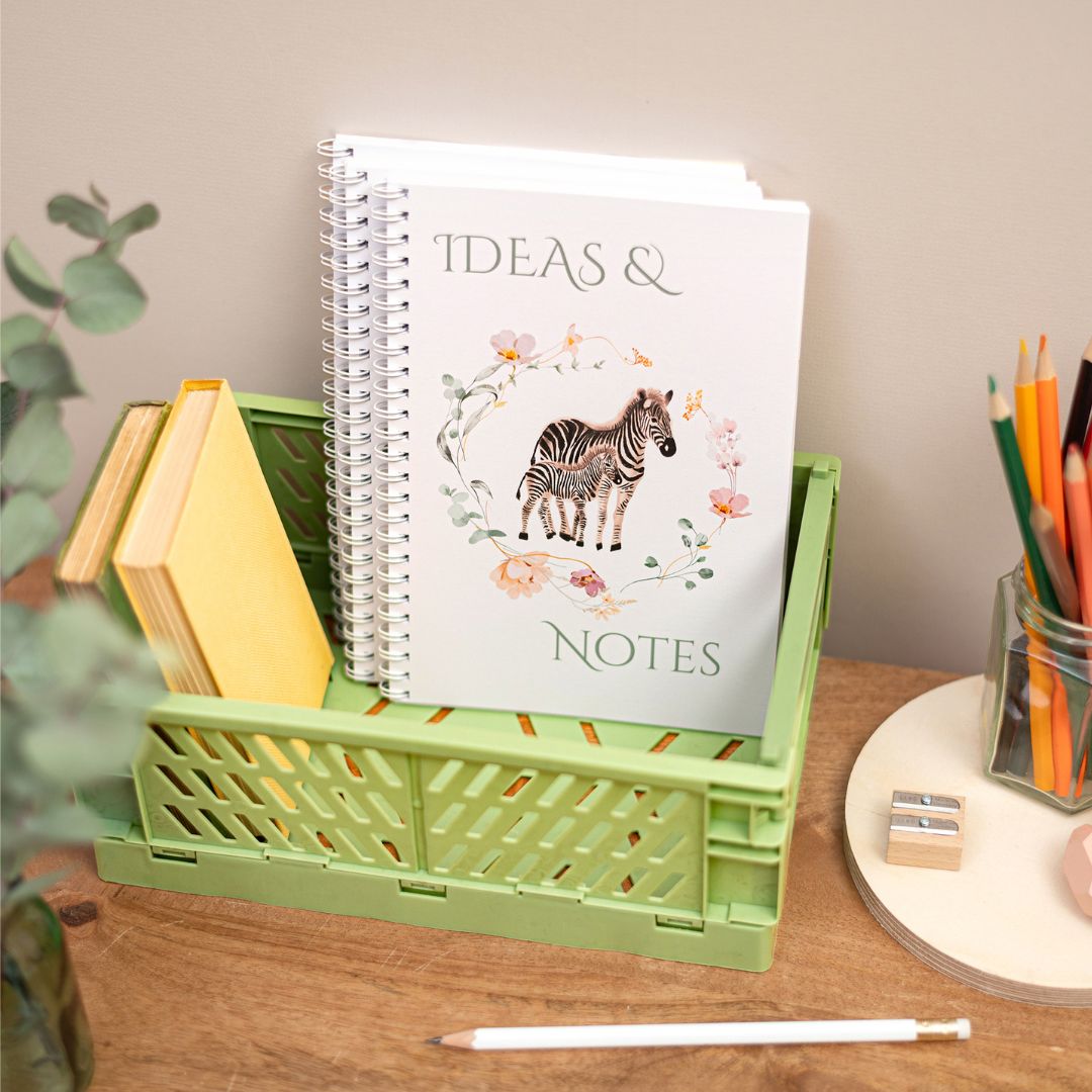 A5 Zebra Handmade Wirebound Notebook. A handmade notebook with a zebra design on the cover, titled 'Ideas & Notes', placed in a green basket on a desk alongside books, pencils, and a plant.