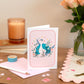A Peacock Blank Everyday card with an elegant peacock design on a soft pink background, displayed on a table with a pink envelope, decorative hearts, and a floral arrangement in the background.