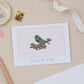 A thank you card with a bird illustration on it, placed on a table with a stamp, ribbon, and other stationery items.