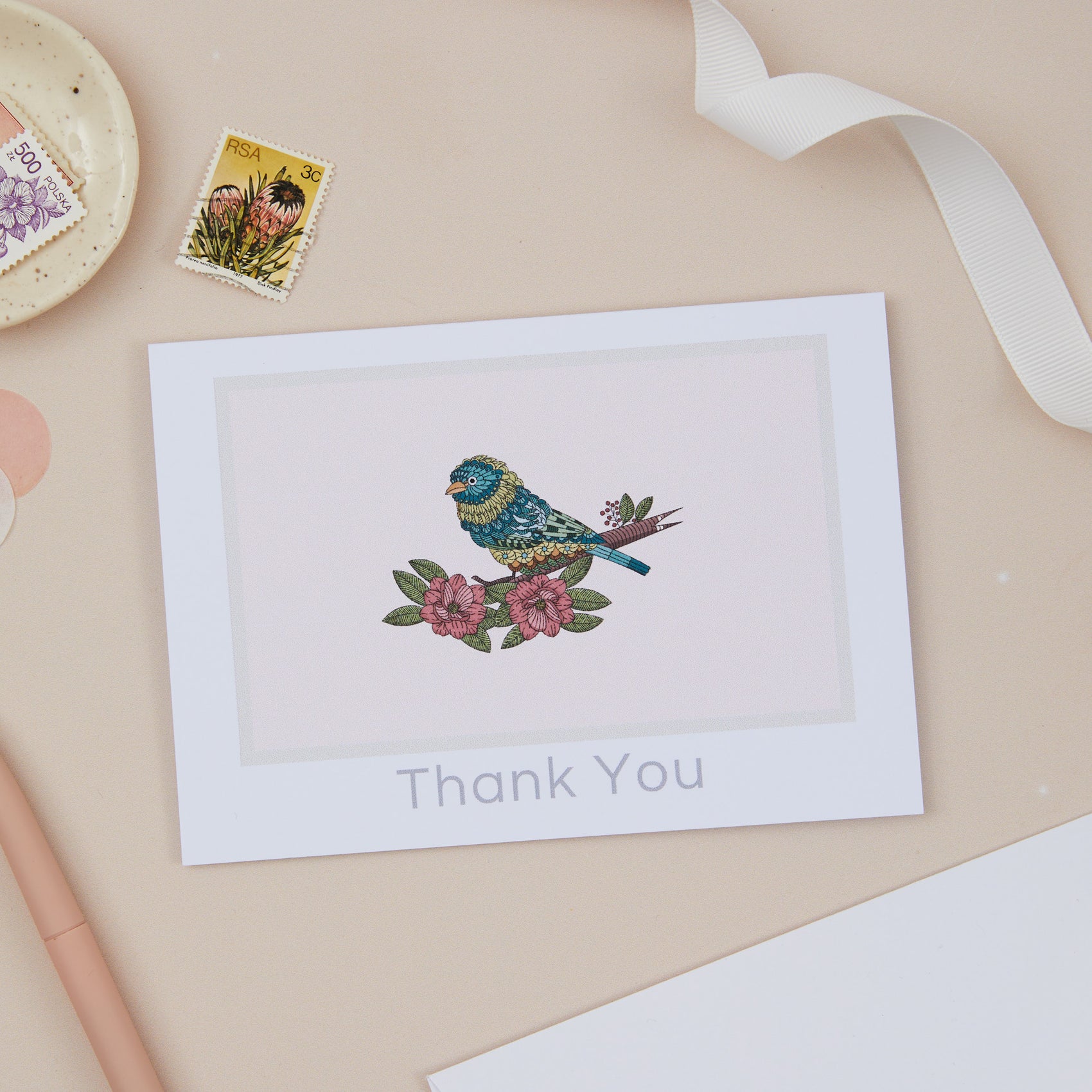 A thank you card with a bird illustration on it, placed on a table with a stamp, ribbon, and other stationery items.