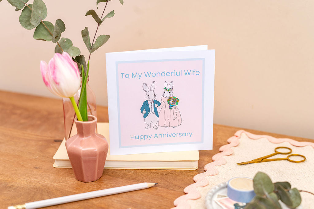 Wife Wedding Anniversary Card with cartoon bunnies and 'Happy Anniversary' text on a wooden surface with flowers and stationery.