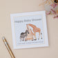 A Baby shower card with animal illustrations on a Mum Zebra & its Baby, Plus a Mum Giraffe and its young. They overlook a Baby Cot. The illustration is set on a pale blue background. The Card is against a neutral background which has a pen and dried flowers
