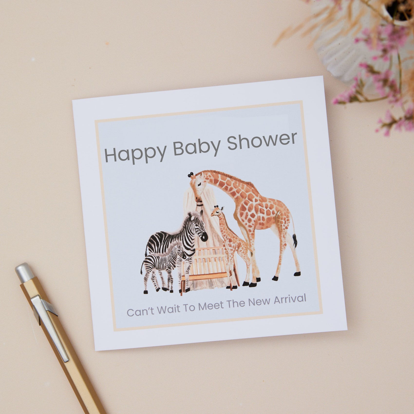 A Baby shower card with animal illustrations on a Mum Zebra & its Baby, Plus a Mum Giraffe and its young. They overlook a Baby Cot. The illustration is set on a pale blue background. The Card is against a neutral background which has a pen and dried flowers