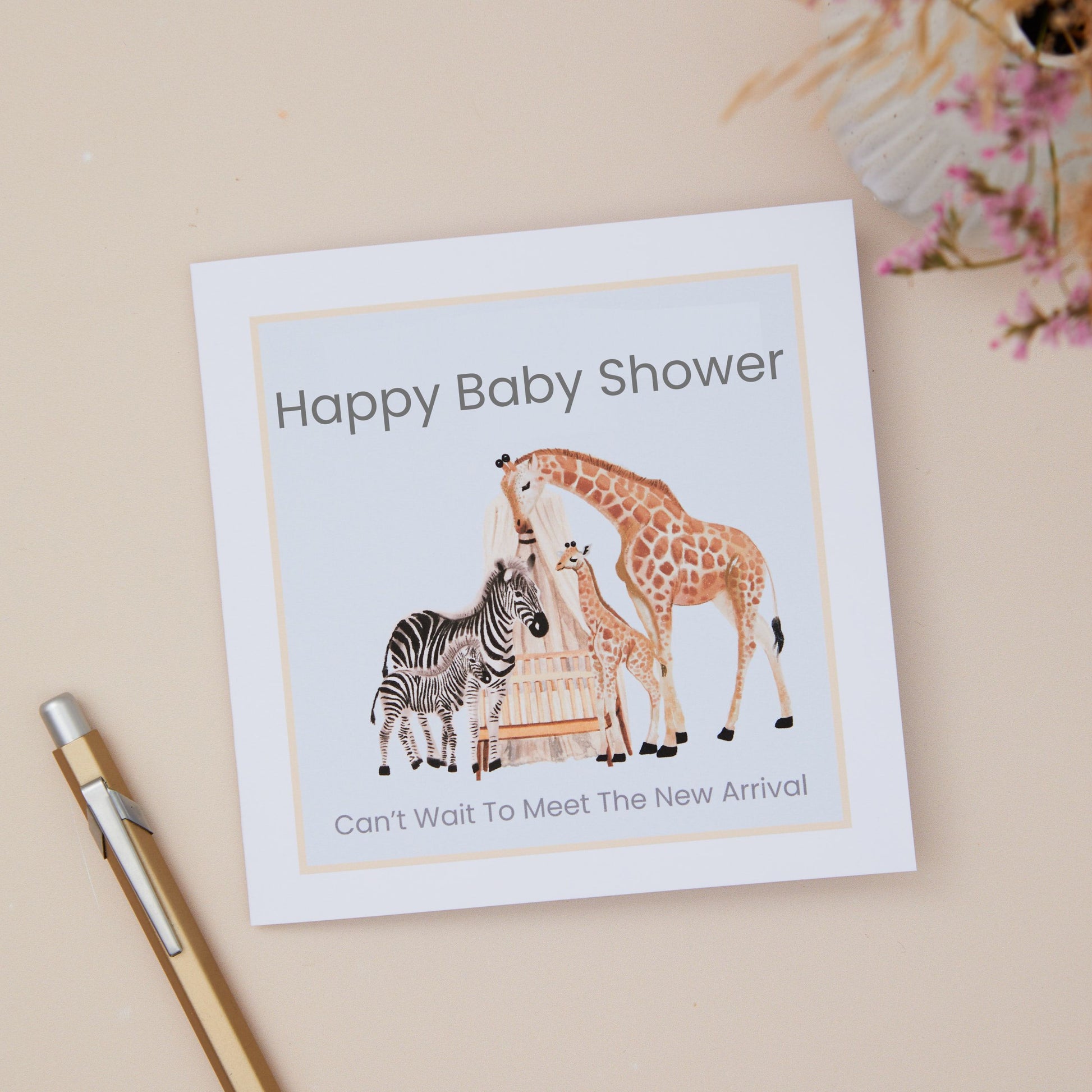 A Baby shower card with animal illustrations on a Mum Zebra & its Baby, Plus a Mum Giraffe and its young. They overlook a Baby Cot. The illustration is set on a pale blue background. The Card is against a neutral background which has a pen and dried flowers