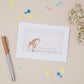 Greeting card featuring a giraffe and it's young on a pink background. The card says - thanks for the gift, and then the name of the mother & Baby bump. The card is set on a neutral background with stationery elements around it.