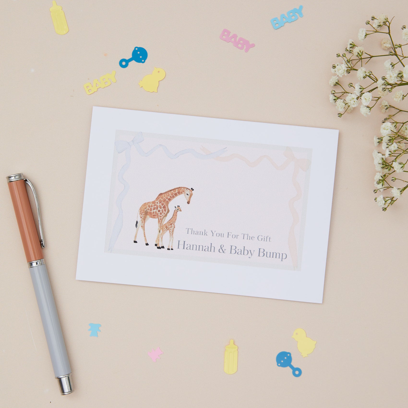 Greeting card featuring a giraffe and it's young on a pink background. The card says - thanks for the gift, and then the name of the mother & Baby bump. The card is set on a neutral background with stationery elements around it.