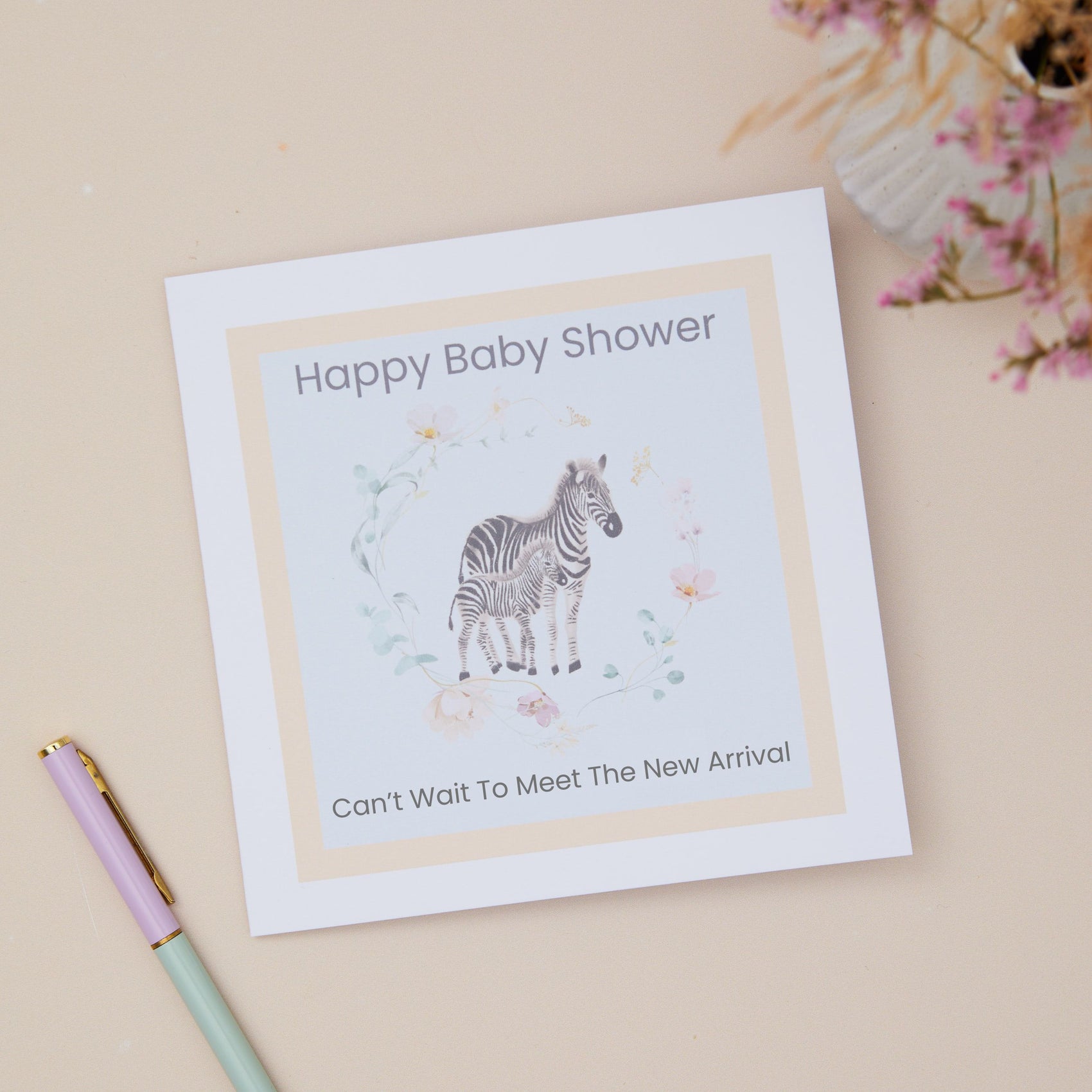 Baby shower card with zebra illustration and text that says happy baby shower, can't wait to meet the new arrival. The cards sits on a beige background