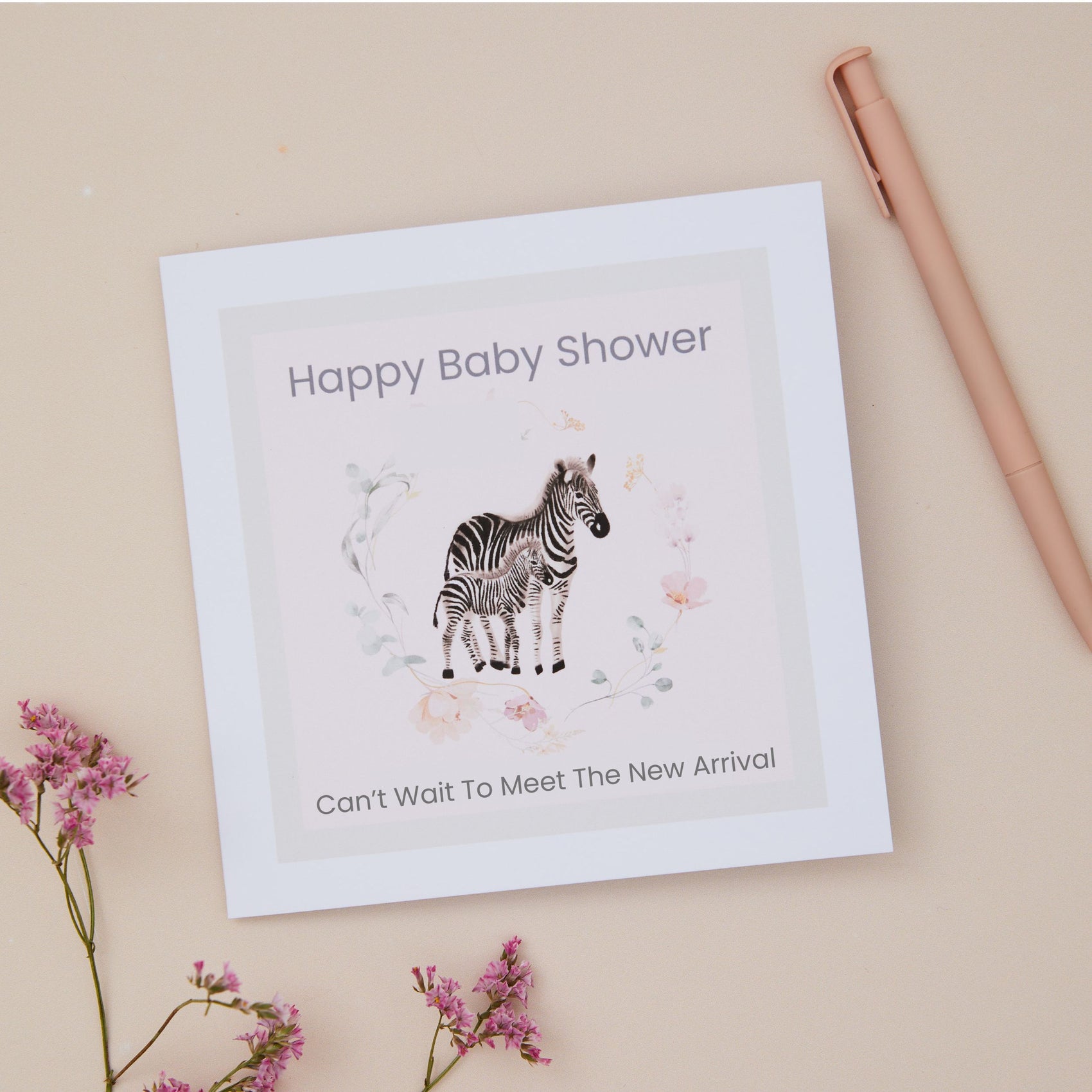 Baby shower card with zebra illustration with a text that sys happy baby shower, can't wait to meet the new arrival. The zebra and floral ring surrounding it sits on a pink background. The card is on a beige background