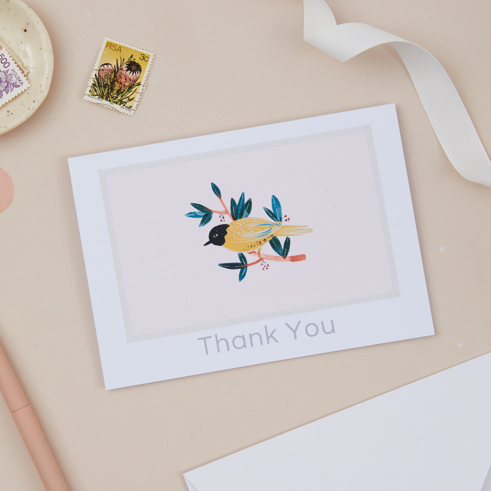 A thank you card with a bird and leaves illustration, placed on a flat surface with a ribbon, stamp, and decorative elements.