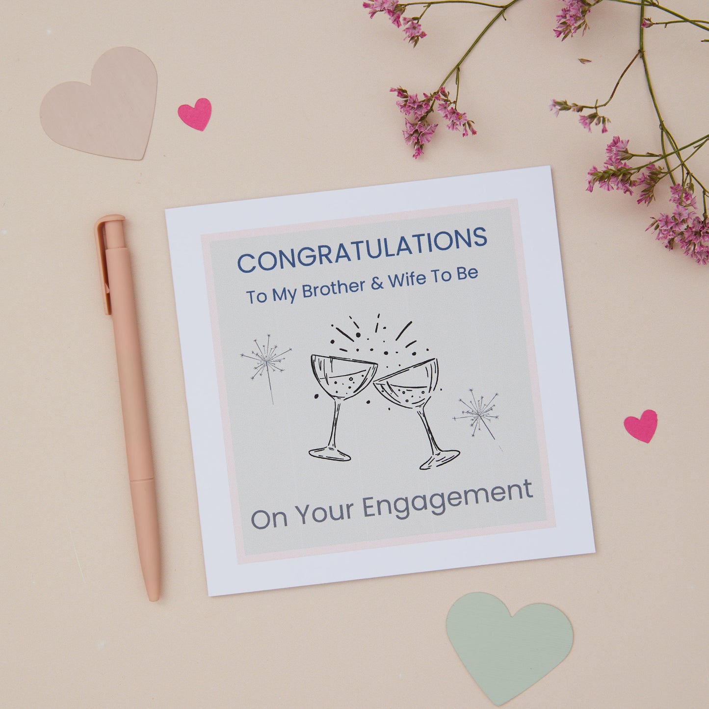 A Brother and wife to be Engagement Congratulations Card featuring two celebratory glasses against a pastel background, with a pink dried flowers bunch, a pink pen, and decorative hearts.