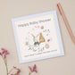 Baby shower card with a bunny sitting in front of Cot. There is a floral delicate ring of flowers around the bunny and the cot. The Bunny and cot are against a pale pink background with a yellow border. The Card sits on a beige background with stationery and dried flowers