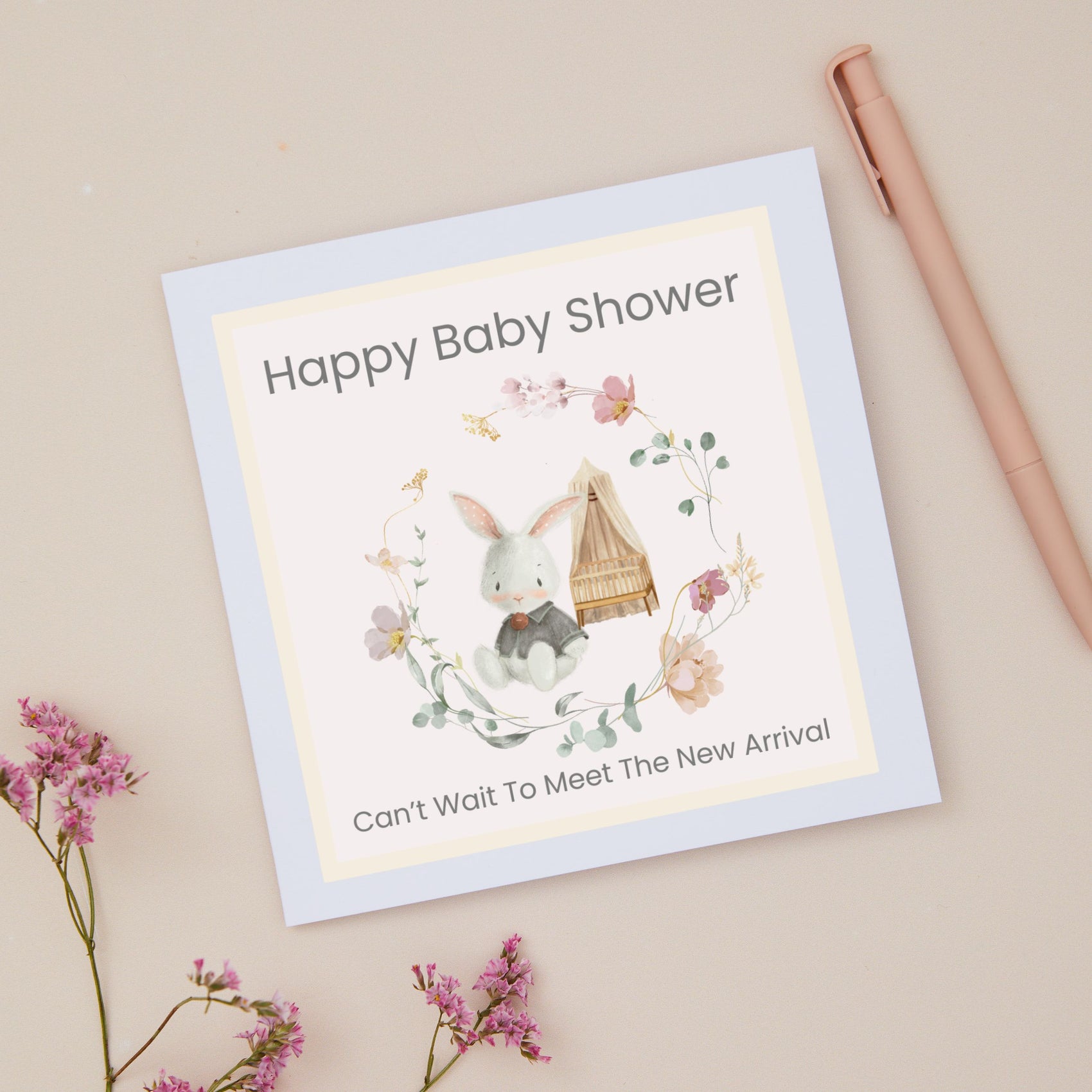 Baby shower card with a bunny sitting in front of Cot. There is a floral delicate ring of flowers around the bunny and the cot. The Bunny and cot are against a pale pink background with a yellow border. The Card sits on a beige background with stationery and dried flowers
