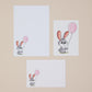 Bunny Rabbit Letter Writing Set showing three illustrations of a cartoon rabbit with pink balloons, Writing Paper, Envelopes and postcards on a white background, displayed on a beige surface.