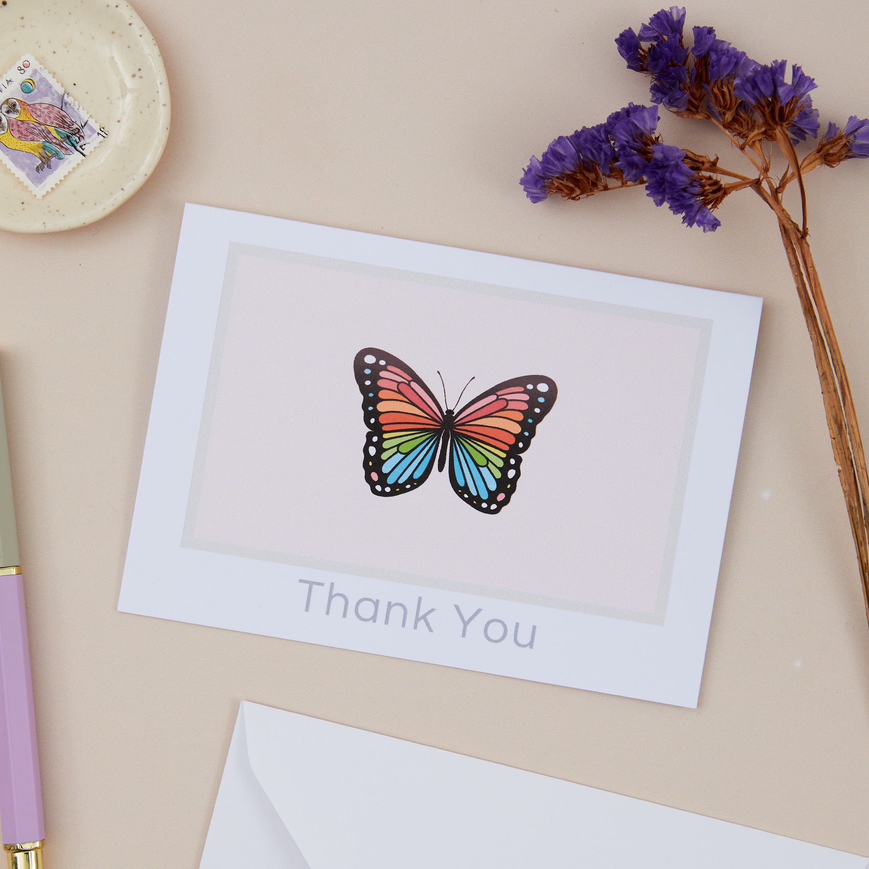 Butterfly Thank You Greeting Card. Butterfly is set against a pink background. The Card is on a beige background with stamps, stationery, and a dried purple foliage.