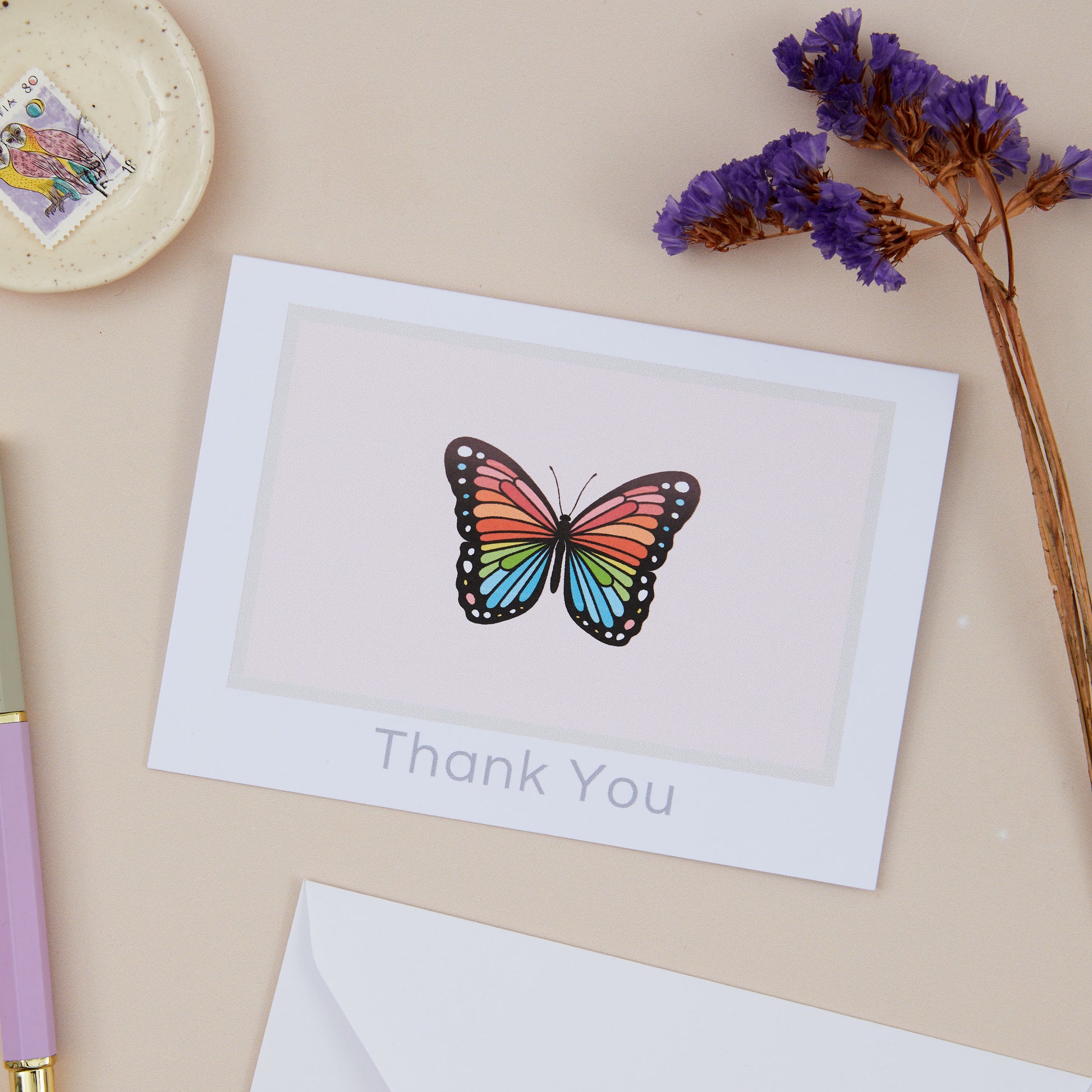 Butterfly Thank You Greeting Card. Butterfly is set against a pink background. The Card is on a beige background with stamps, stationery, and a dried purple foliage.