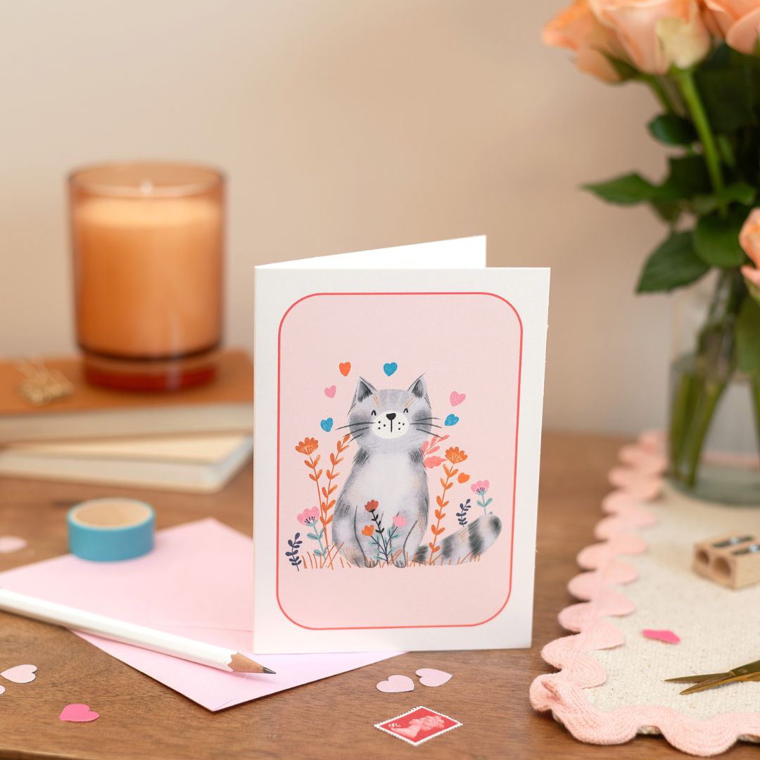 Cat Blank Greeting Card Set of 8 with a cute cat illustration on a pink background, displayed on a desk next to a pink envelope, with a candle and flowers in the background.