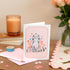 Cat Blank Greeting Card Set of 8 with a cute cat illustration on a pink background, displayed on a desk next to a pink envelope, with a candle and flowers in the background.