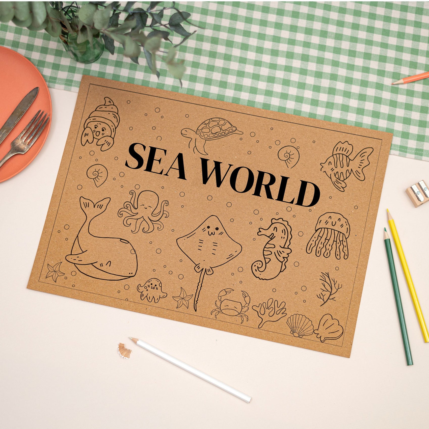 Children's Seaworld Colouring in Placemat. The Colouring mat features cartoon style seaworld characters, and has the text of 'Sea World' on the front. The Mat sits on a checkered tablecloth