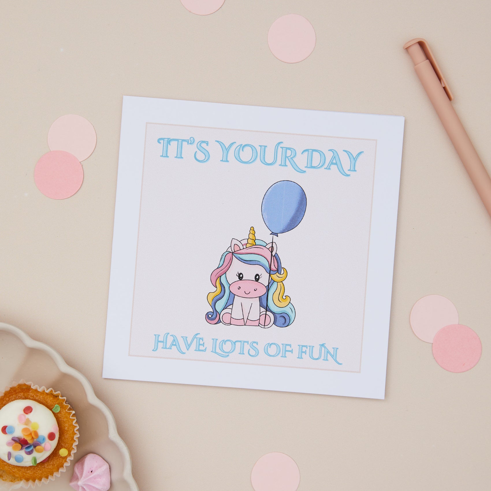 Children's Unicorn Celebration Card Baby Unicorn holding a balloon with the caption it's your day have lots of fun. The card is set again a neutral background with stationery and party cup cakes