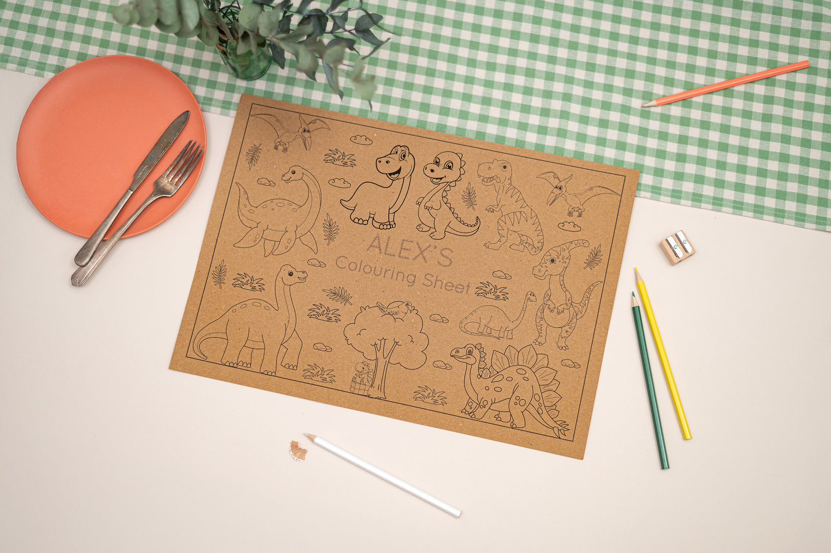 Kids Dinosaur colouring Mat. Mat illustrates different types of cartoon style dinosaurs. The Colouring mat is made with Kraft Brown Eco Friendly paper. The Mat is on a table with coloring supplies