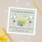 Congratulations on passing exam card with an illustration of a pile of books and multi coloured balloons on either side. there is a star burst of sprinkles across the card