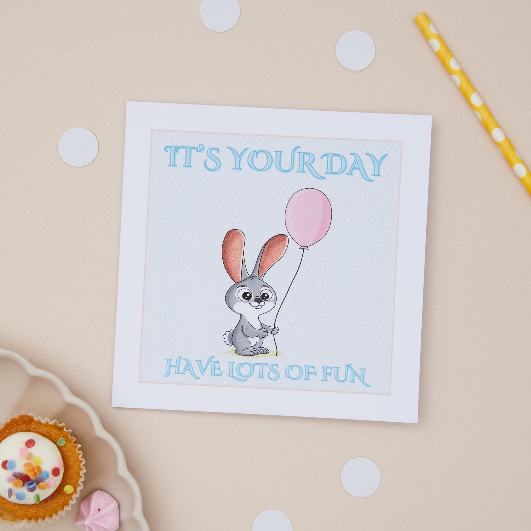 Kids Birthday Greeting card with a cartoon rabbit holding a pink balloon, with the text 'IT'S YOUR DAY HAVE LOTS OF FUN' on a 7x7 inch format, displayed on a neutral background with polka dots and a cupcake.
