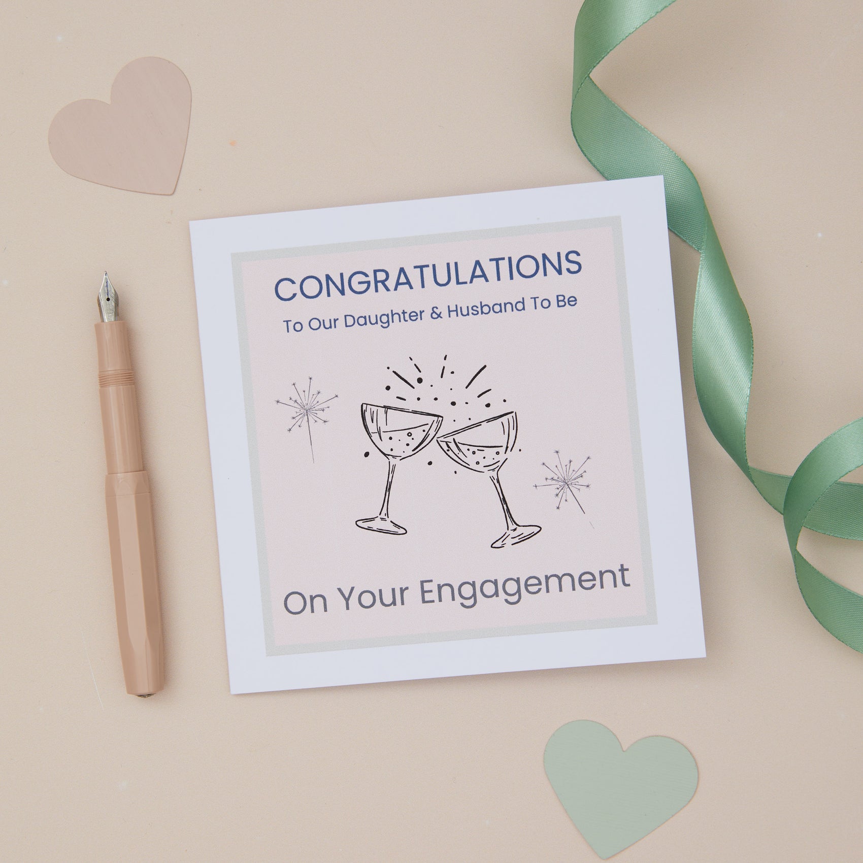 A Daughter Engagement Congratulations Card with two celebratory glasses design, accompanied by a beige pen, a green decorative ribbon running alongside it, and decorative heart-shaped cutouts on a beige background.