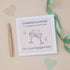 A Daughter Engagement Congratulations Card with two celebratory glasses design, accompanied by a beige pen, a green decorative ribbon running alongside it, and decorative heart-shaped cutouts on a beige background.