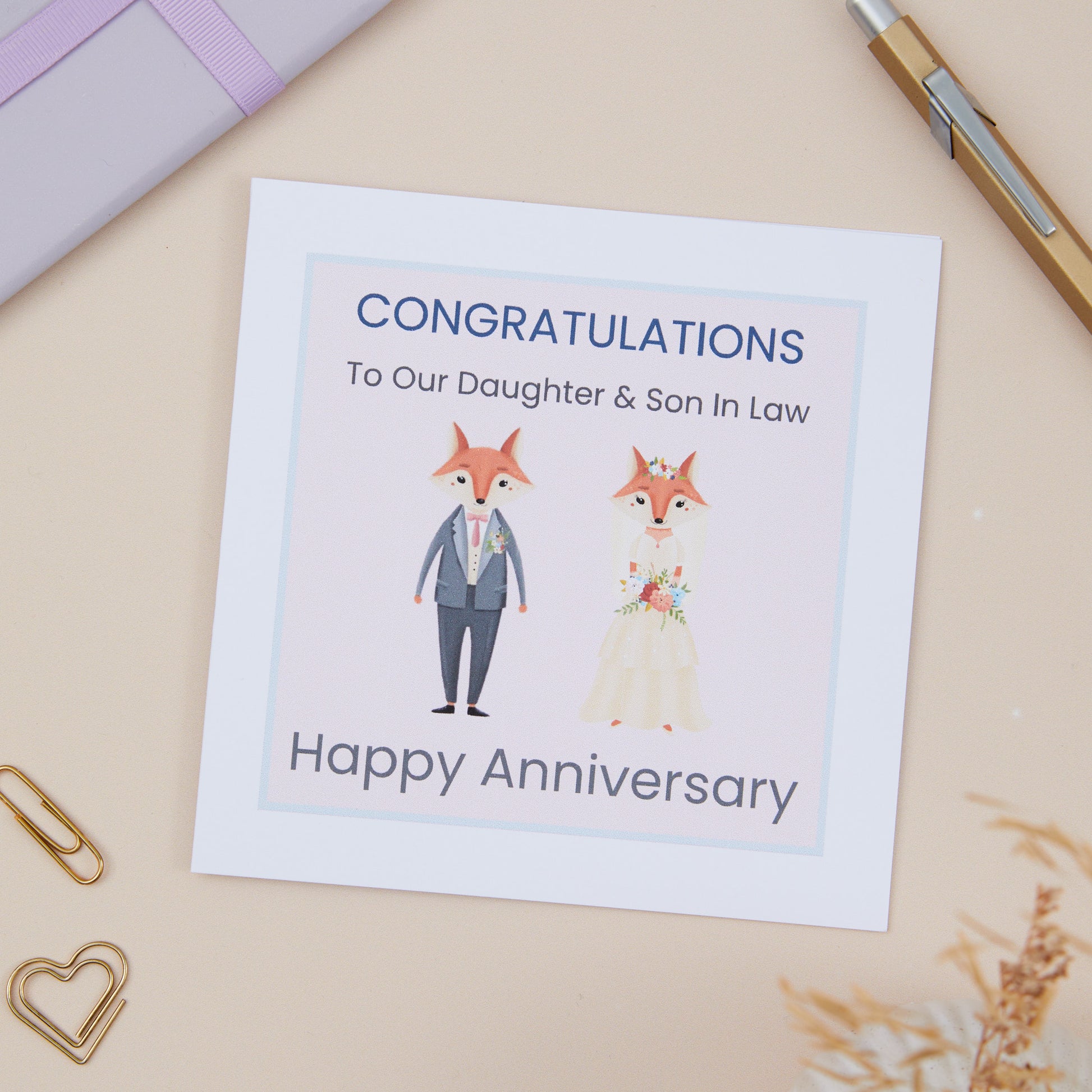 A Daughter Son In Law Wedding Anniversary Card with an illustration of a fox couple dressed in wedding attire, with text that reads 'Congratulations to our Daughter & Son-in-Law, Happy Anniversary'. The card is placed on a neutral background with a white envelope and some decorative elements like a heart clip and dried flowers.