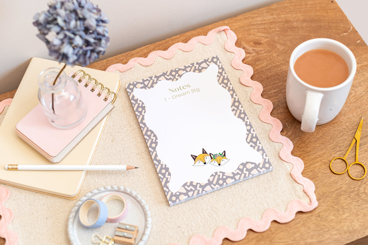 An A5 daily planner with a colorful fox design and a 'Dream Big' motif on the cover, displayed on a wooden desk alongside a cup of tea, notepad, and stationery items.