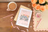 Personalised A5 Notebook which is wire bound. Features two elephants on the front against a pink background. The notebook is on a wooden table with a cup of coffee, flowers, and stationery items.