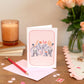 Elephant Greeting Cards Set of 8 with a design of two elephants facing each other on a soft pink background, accompanied by a matching pink envelope, a pen, and some decorative hearts on a table.