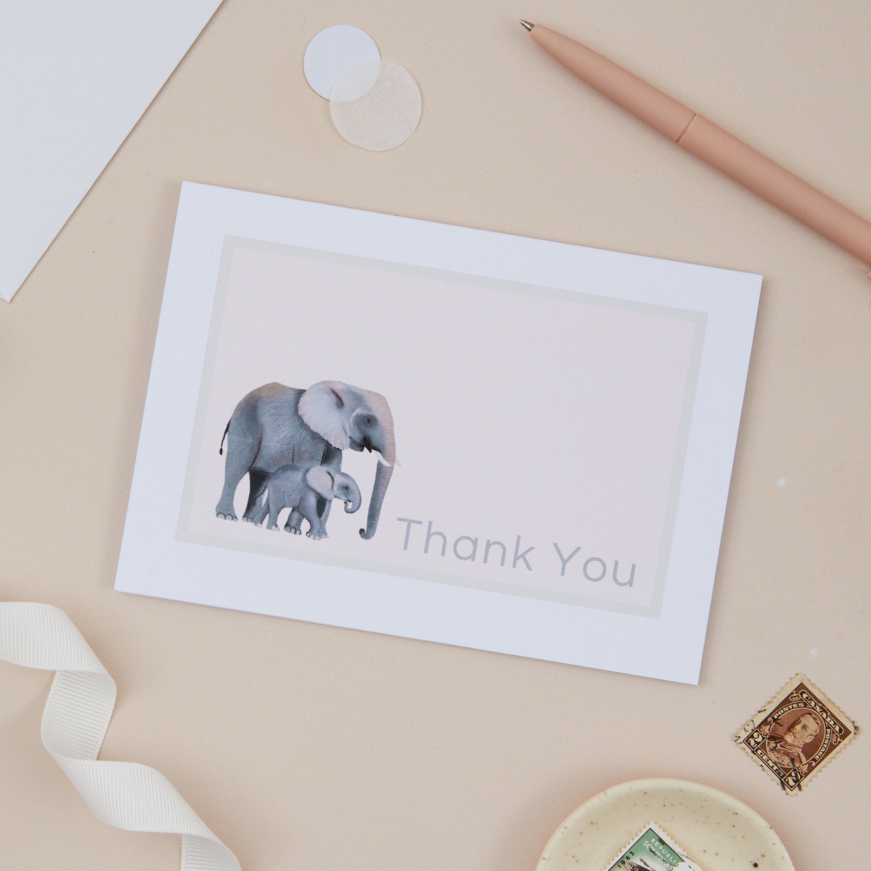 A thank you greeting card with an elephant design, accompanied by a luxury white lined envelope, a pen, ribbon, and decorative items.